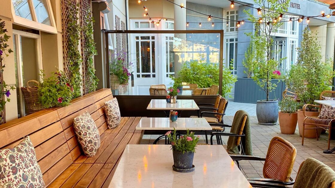 Cosy hotel terrace with wooden benches, soft cushions, string lights, and lush potted plants, perfect for relaxing outdoors.