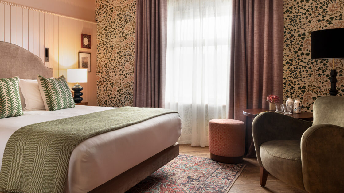 Cosy hotel room with plush bed, patterned cushions and elegant decor, featuring a green throw, vintage rug, and comfortable seating.
