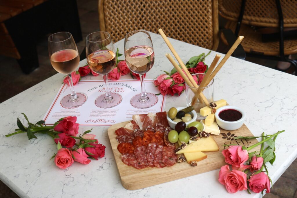 A charcuterie board with meats, cheeses, olives, walnuts, and breadsticks, three glasses of rosé, and pink roses on a white table with a wicker chair in the background.