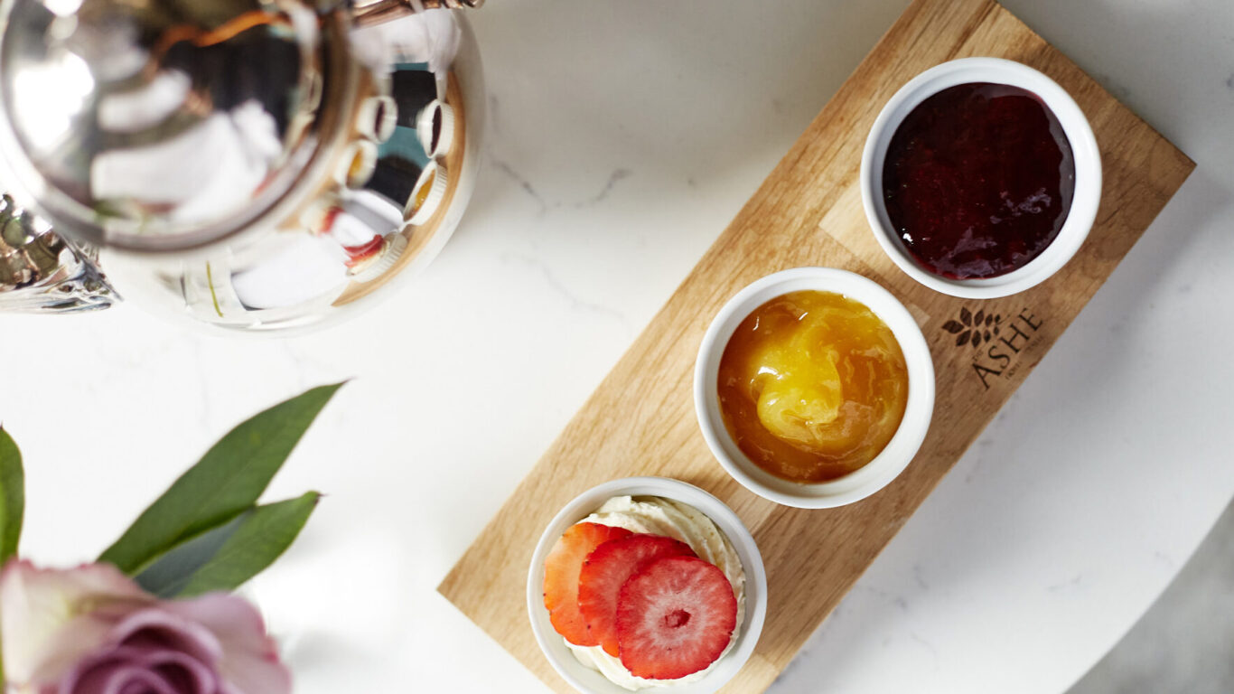 Three small bowls on a wooden tray with clotted cream topped with strawberries, yellow marmalade, and red jam, next to a silver teapot and a pink rose on a white table.