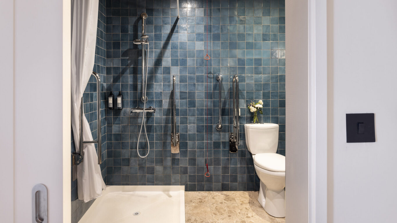 Accessible bathroom with roll-in shower, grab rails, blue tiles, and fresh flowers, offering comfort and convenience.